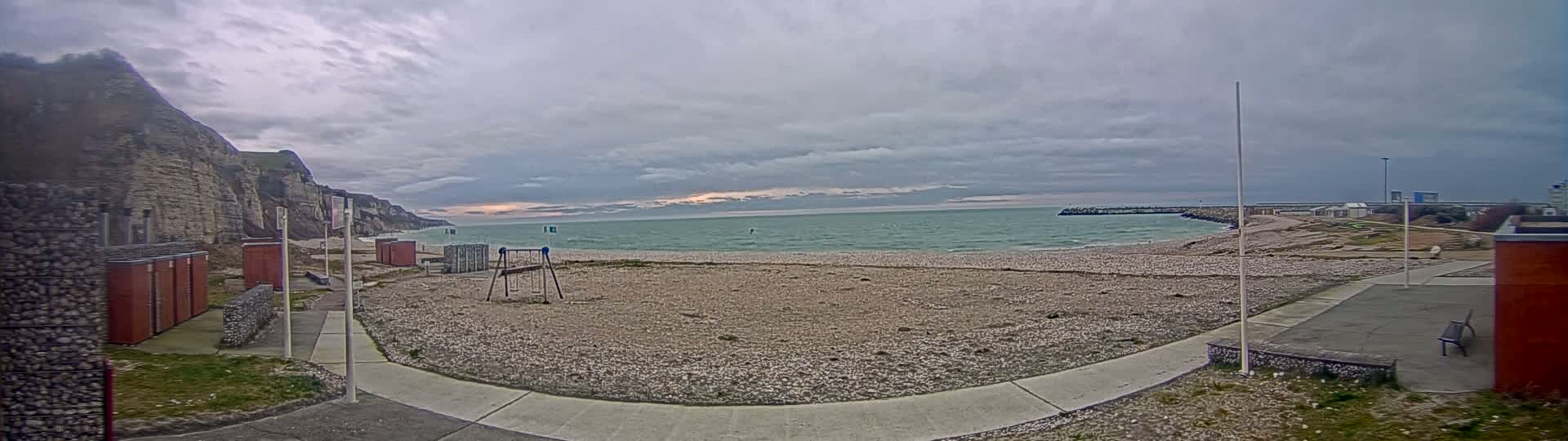A pebble beach stretches towards a calm sea under a cloudy sky, backed by a cliff face and a small playground.