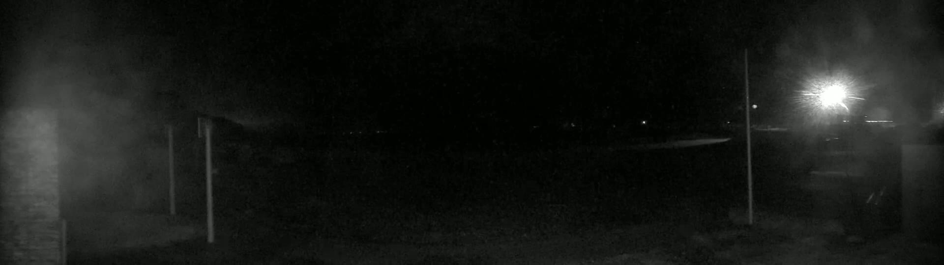 A dark, nighttime scene shows a mostly empty paved area, illuminated by a bright light source in the distance, with hazy or foggy conditions present.