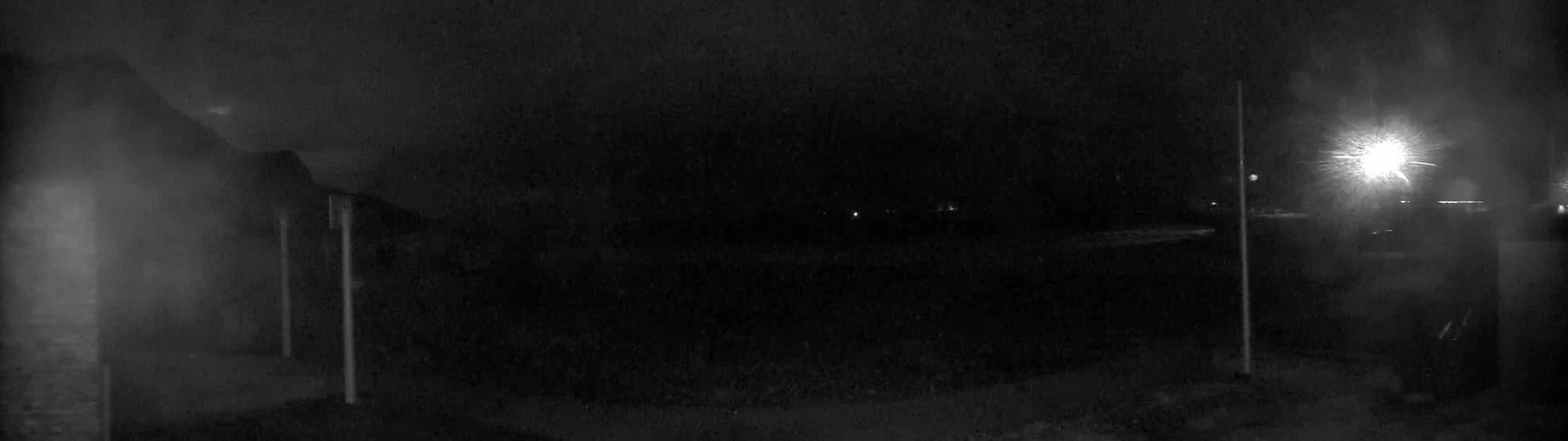 A nighttime, blurry, low-light image shows a dark, possibly snowy or foggy, outdoor scene with indistinct shapes and two tall, thin poles, and a bright light source in the distance.