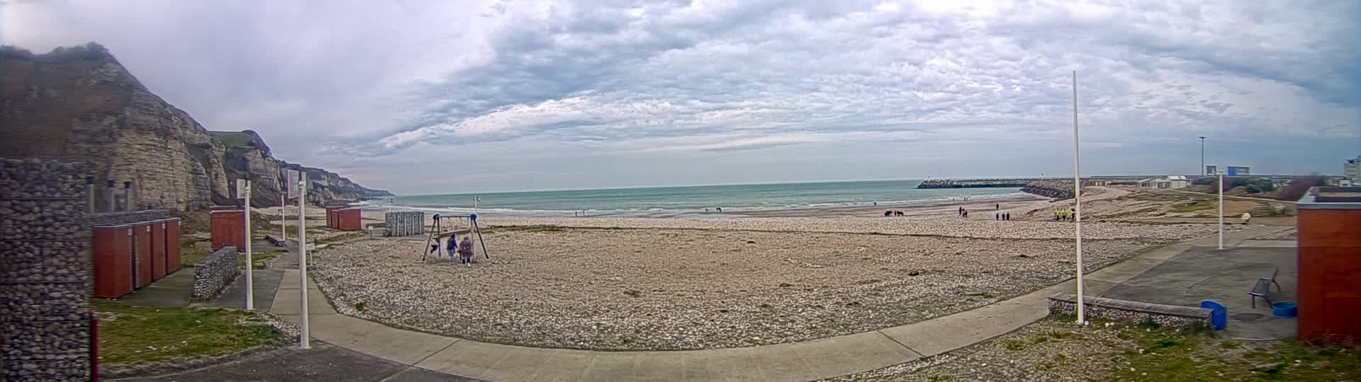 A mostly cloudy day at a pebble beach with a few people, a playground, and a rocky cliff in the background.