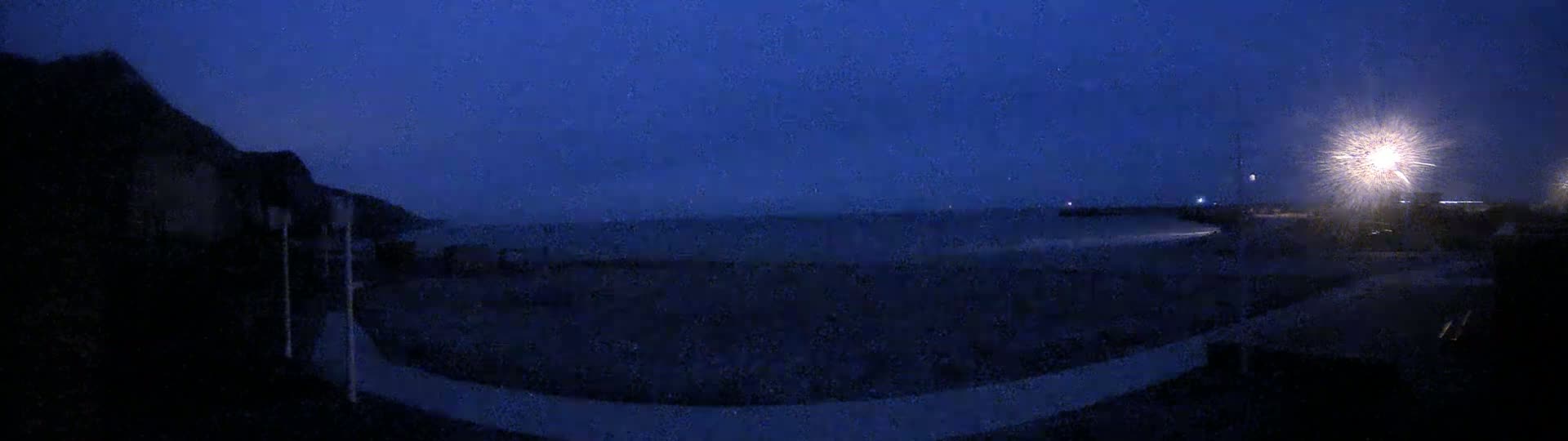 A dark, nighttime coastal scene shows a dark body of water under a dim, possibly stormy sky, with lights visible in the distance along the shore.