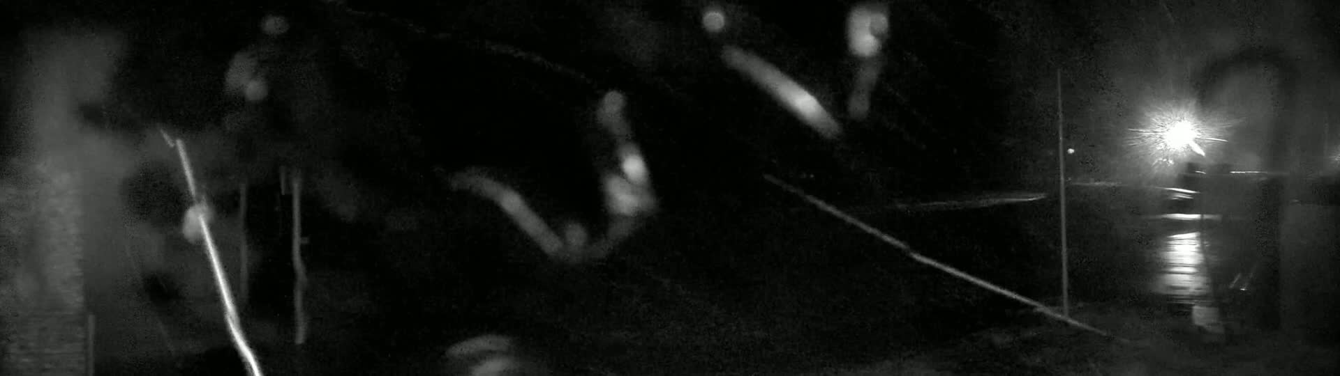 Blurry figures walk at night in the rain, illuminated by a distant light source.