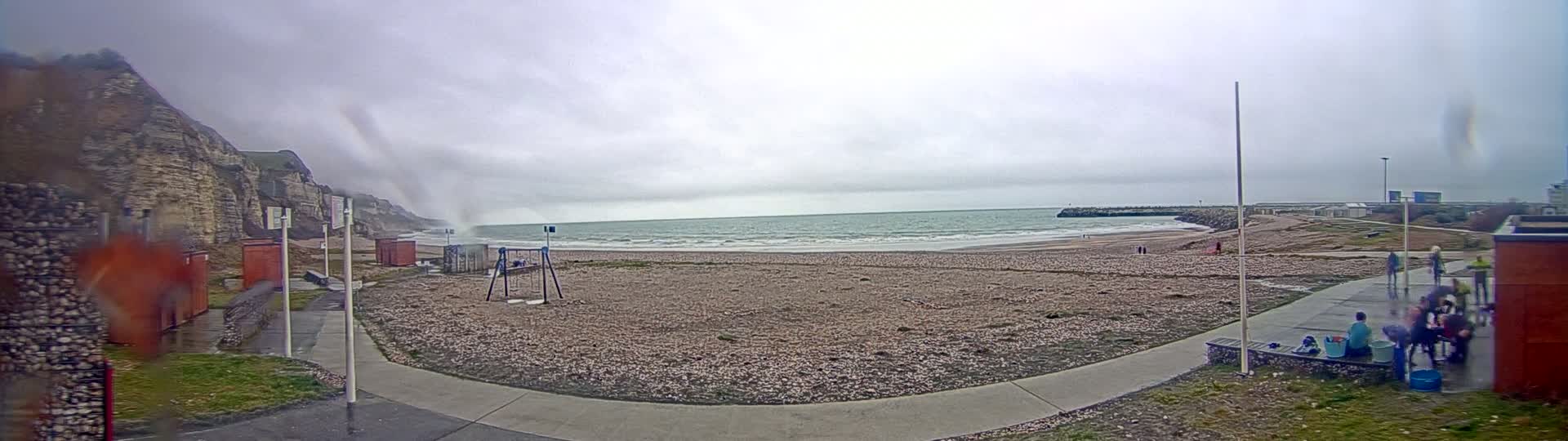 A pebble beach with a few people on it is seen under an overcast sky, with a rocky cliff to the left and a calm sea in the distance.