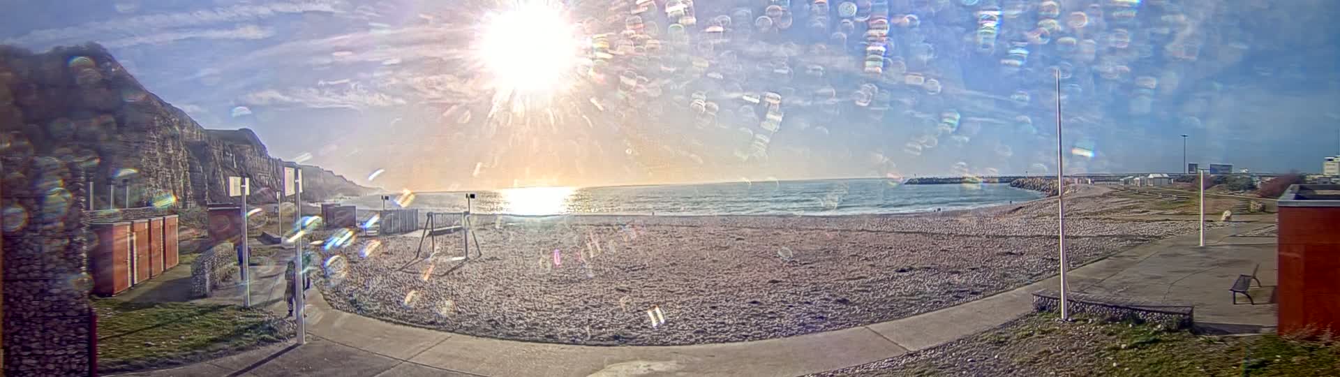 A mostly sunny day at a pebble beach with a view of a cliff face and calm ocean waves.
