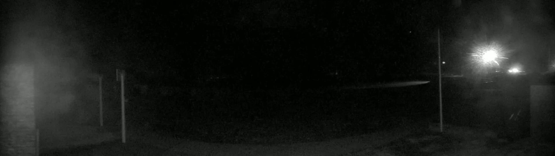A nighttime outdoor scene shows a dimly lit area with some posts and bright lights in the distance, under dark and possibly foggy conditions.