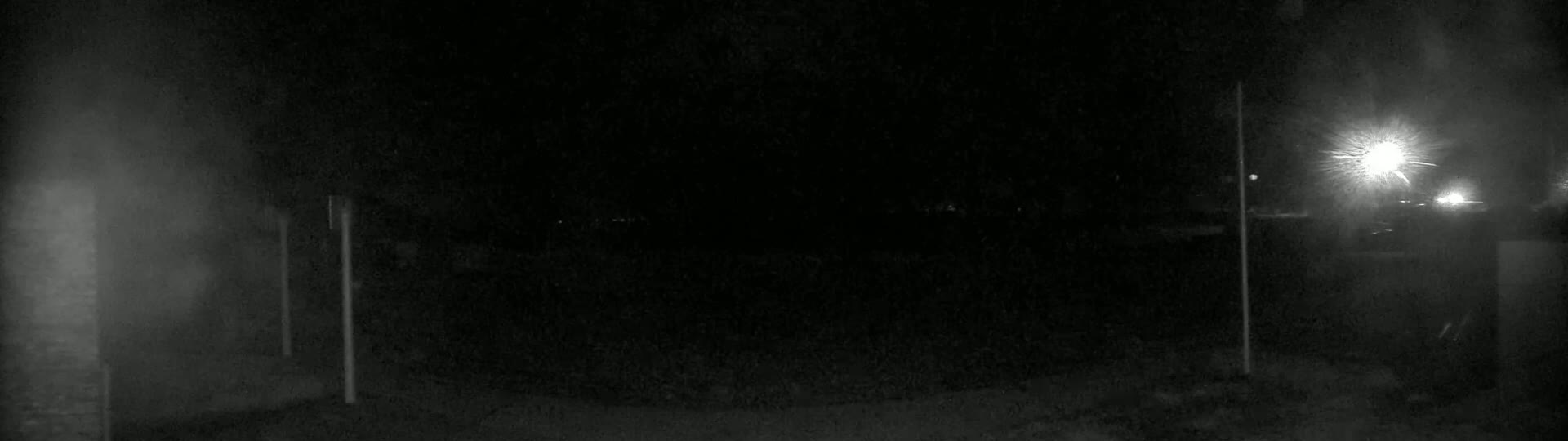 A nighttime, dimly lit outdoor scene shows a dark, mostly empty expanse of ground with two vertical poles and a bright light source in the distance.