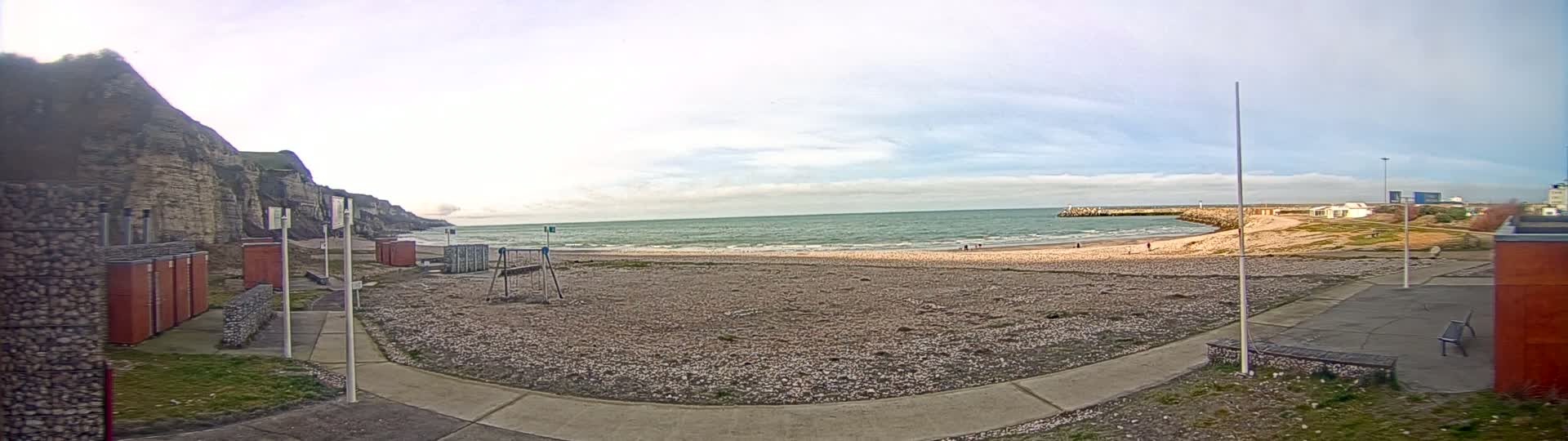 A mostly cloudy day reveals a pebble beach with a calm sea, backed by a tall cliff and some small buildings.