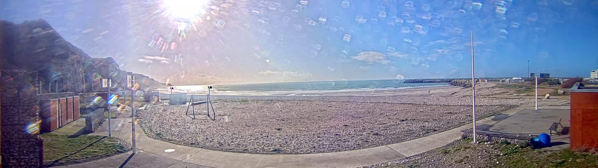 A sunny, slightly hazy day at a beach with a rocky cliff in the background, showing a sandy beach with a walkway and buildings nearby.