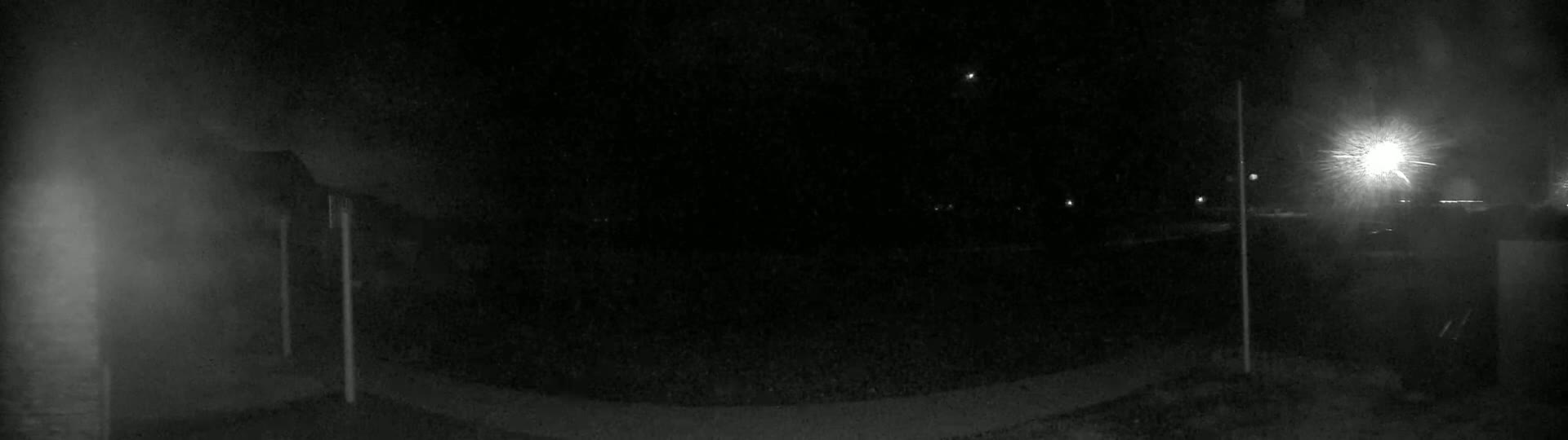A nighttime, low-light image shows a dark, outdoor area with a few distant lights and two vertical poles in the foreground.