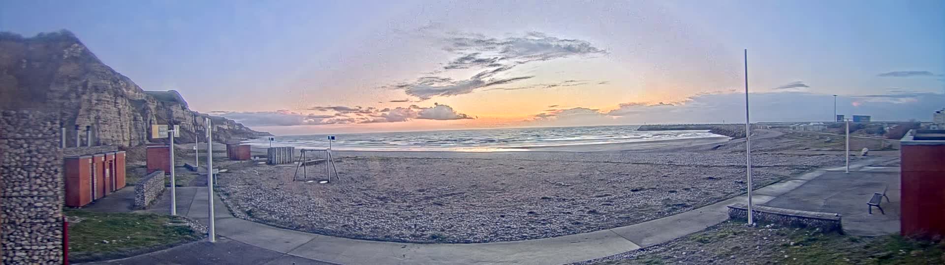 A mostly empty pebble beach at sunset, with a rocky cliff to the left and a breakwater to the right, under a partly cloudy sky.
