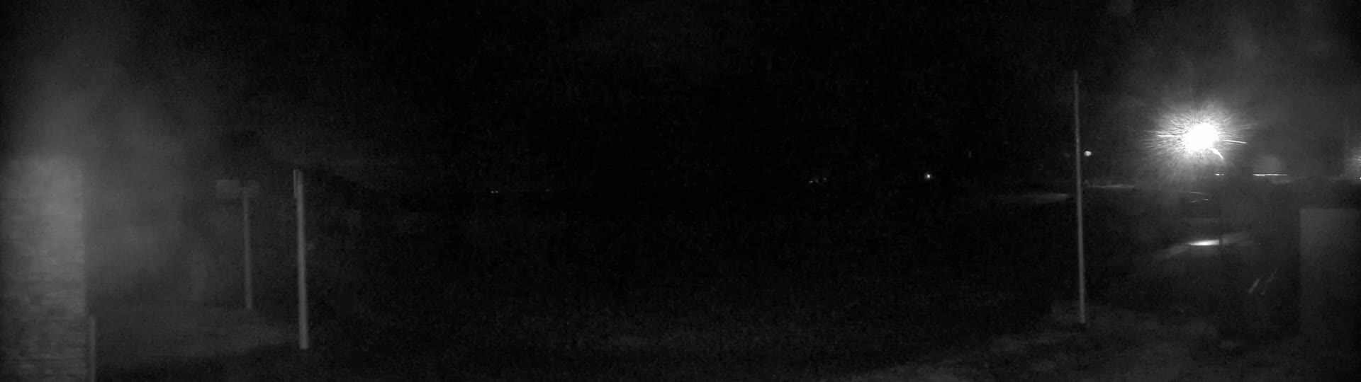 A dimly lit nighttime scene shows a dark, snowy ground with two tall, thin posts and a bright streetlight in the distance.