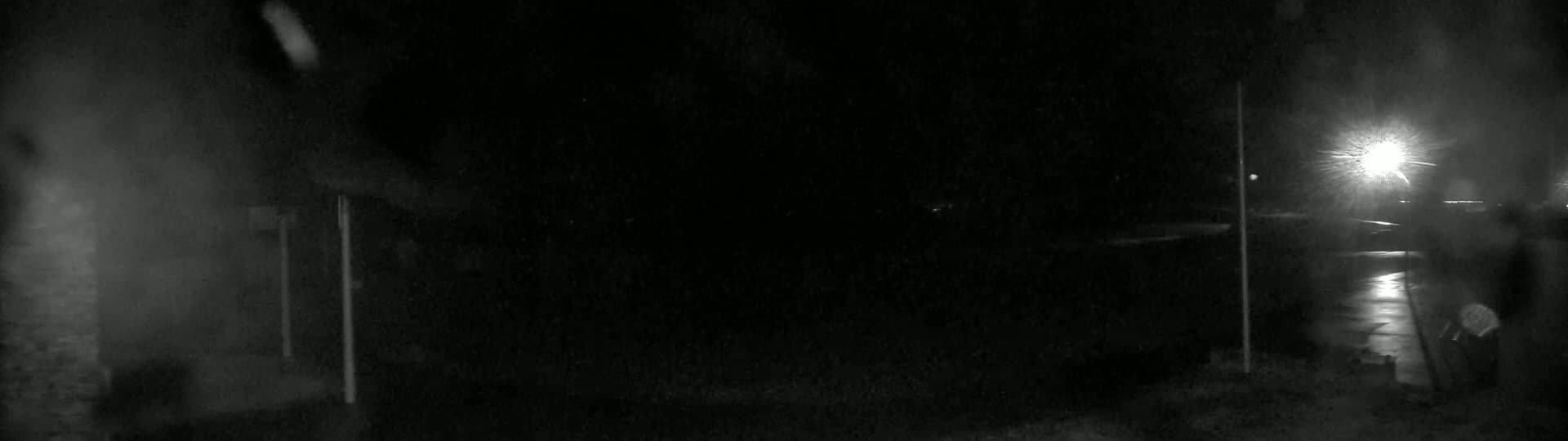 A dark, nighttime scene shows a paved area illuminated by a bright light source in the distance, with light rain or snow falling.
