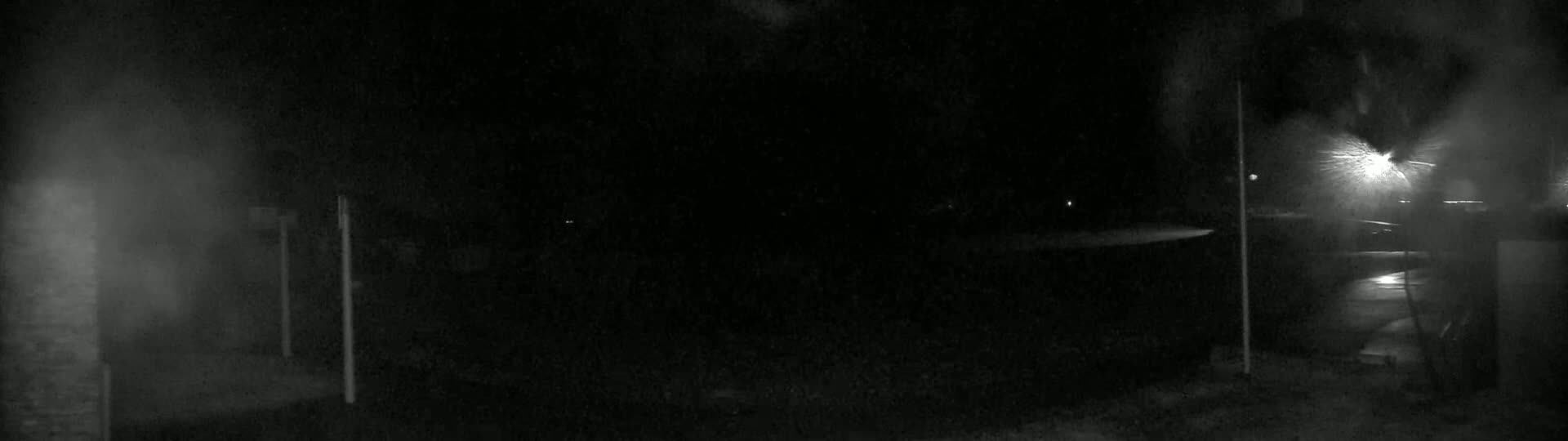 A dark, nighttime scene shows a dimly lit area with two tall, thin posts, possibly light posts, and a wet ground.