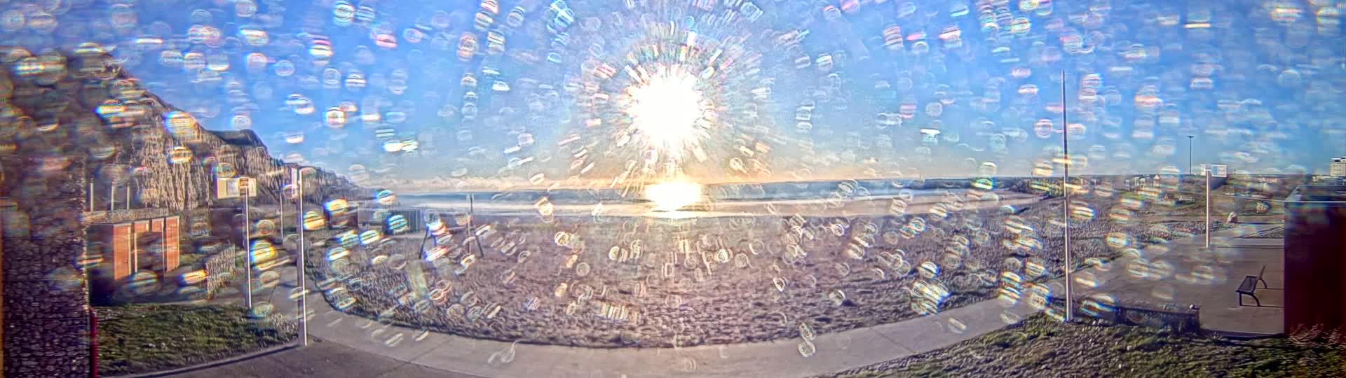 A bright, sun-filled beach scene is viewed through a rain-covered window, showing a sandy beach, ocean, and buildings in the background.