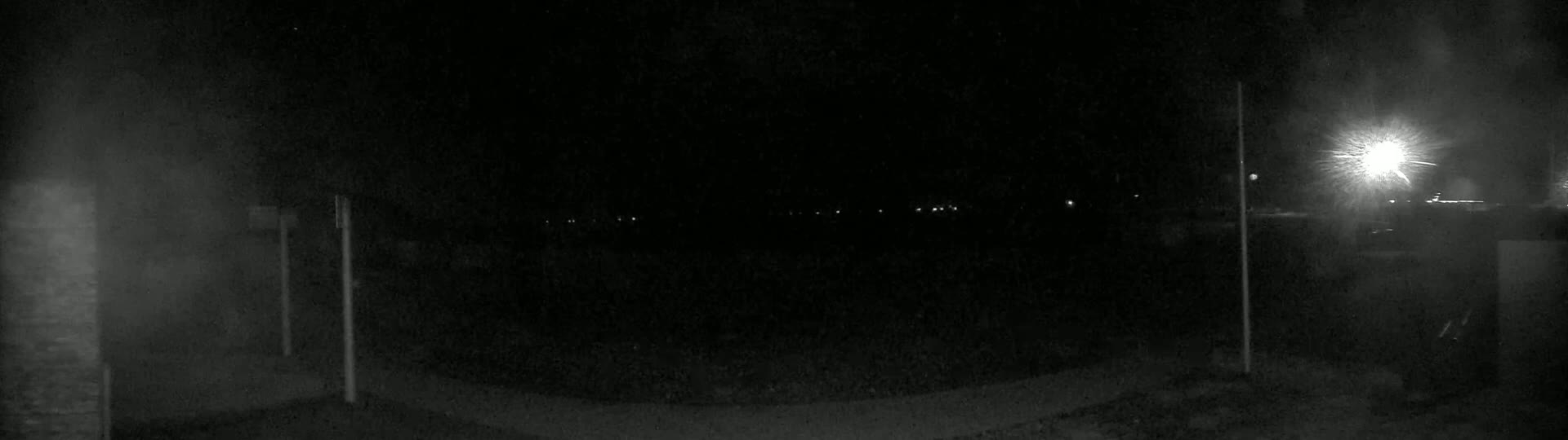 A dark, nighttime scene shows a paved area with faint lights in the distance and tall, thin posts on either side of the frame.