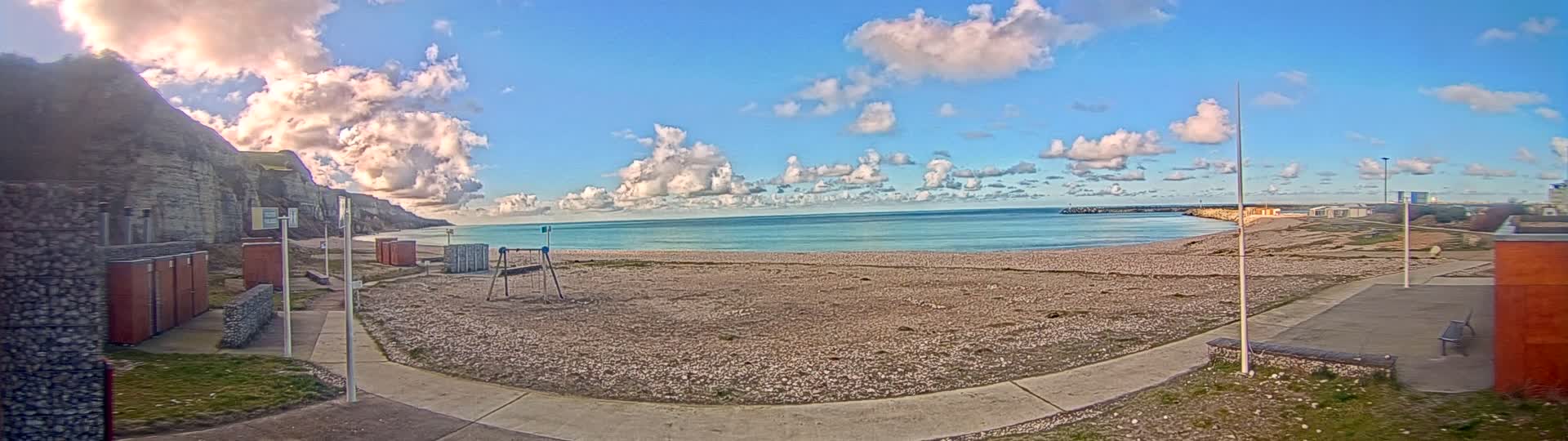 A mostly sunny day shows a pebble beach with a calm turquoise sea, white buildings, and a rocky cliff face under a partly cloudy sky.