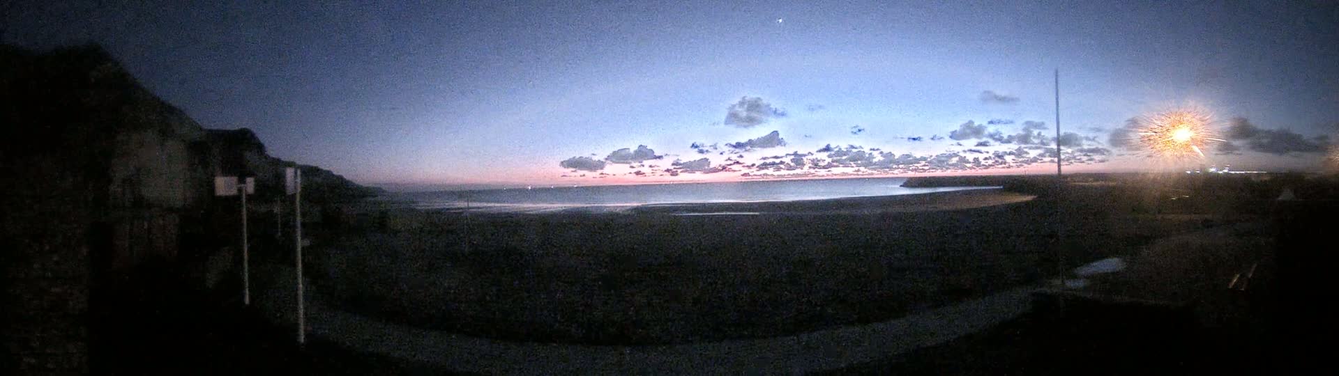 A dark, rocky shoreline at twilight overlooks a calm sea with a pink and purple sunset behind a thin layer of clouds.