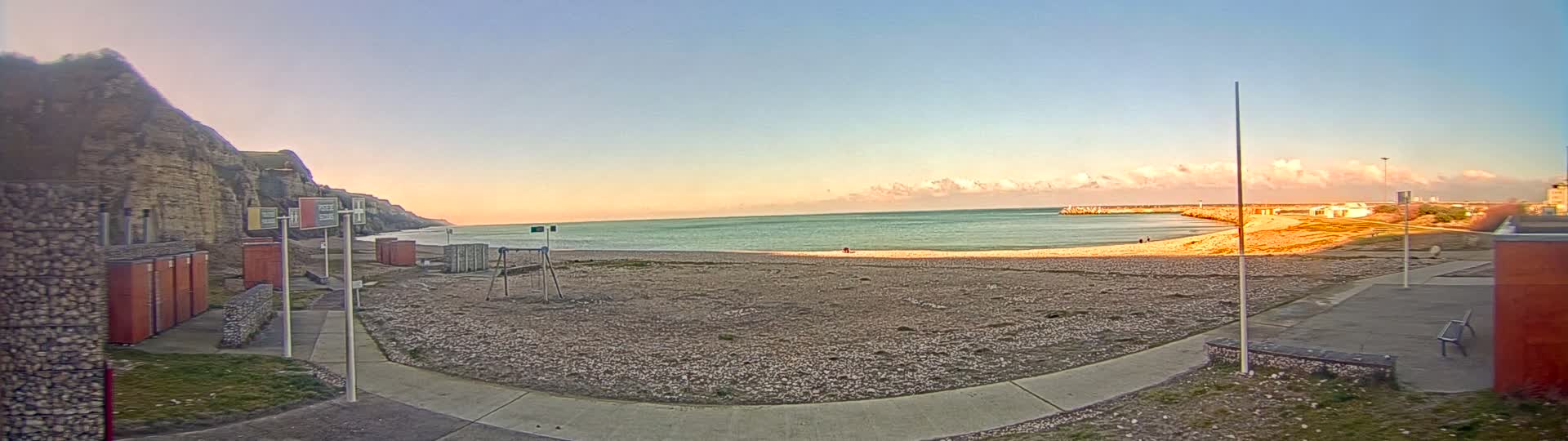 A pebble beach with calm, light-green water under a mostly clear sky with some pink clouds at the horizon.