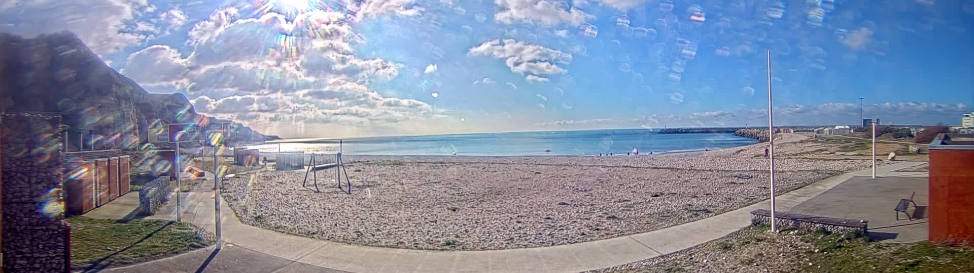 A sunny day with partly cloudy skies shows a sandy beach next to a calm sea with a rocky cliff in the background.
