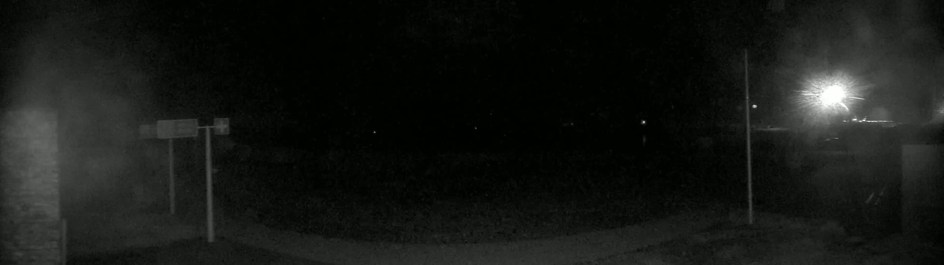 A nighttime, dimly lit outdoor scene shows a dark roadway with two poles and signs, and a bright light source in the upper right corner.