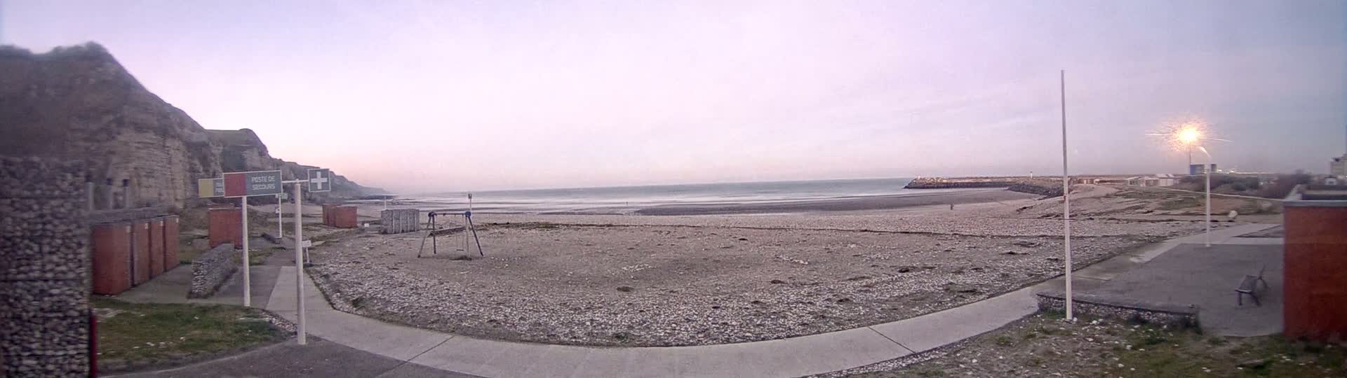 A pebble beach stretches to a calm sea under a pale, dusky sky, backed by a high cliff and a small harbor.