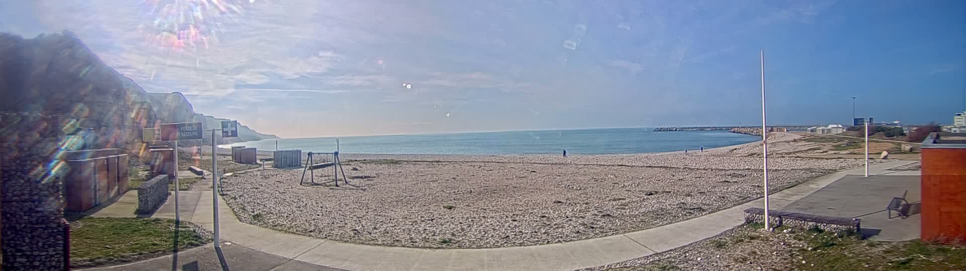 A mostly sunny day at a pebble beach, with a calm sea, a white cliff in the background, and a few people walking on the beach.