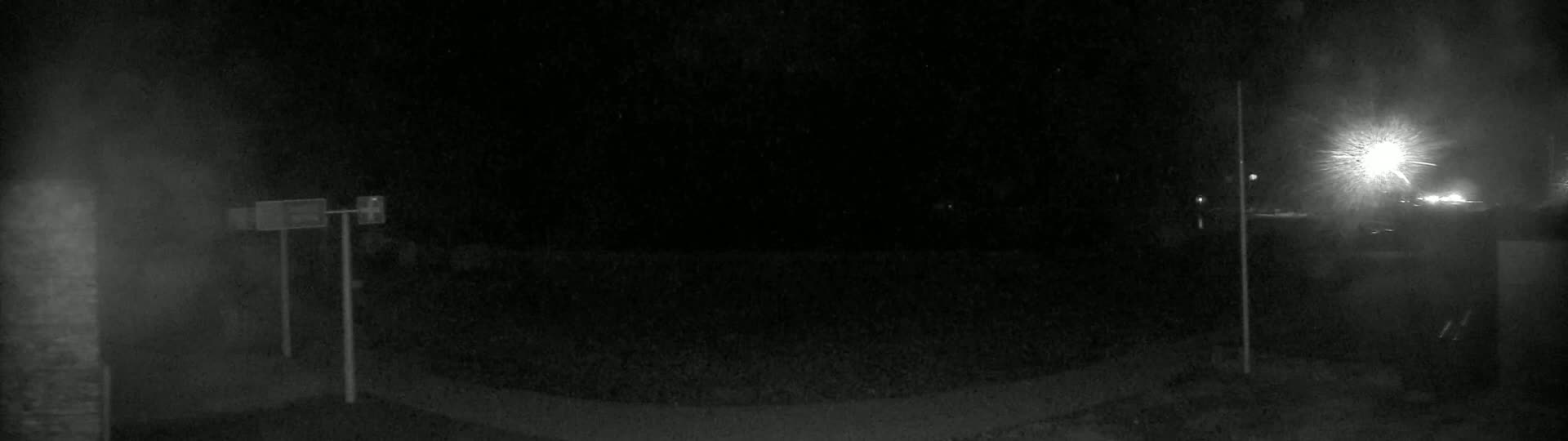 A nighttime, low-light view of a dark, empty roadway with two upright posts and signs visible, illuminated by distant bright lights.