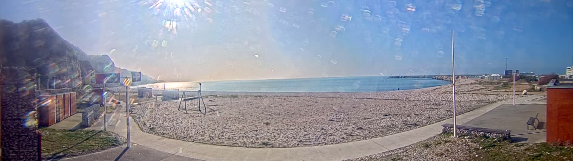 A mostly sunny day at a sandy beach with calm water, a rocky cliff in the background, and a paved walkway leading to a small building.