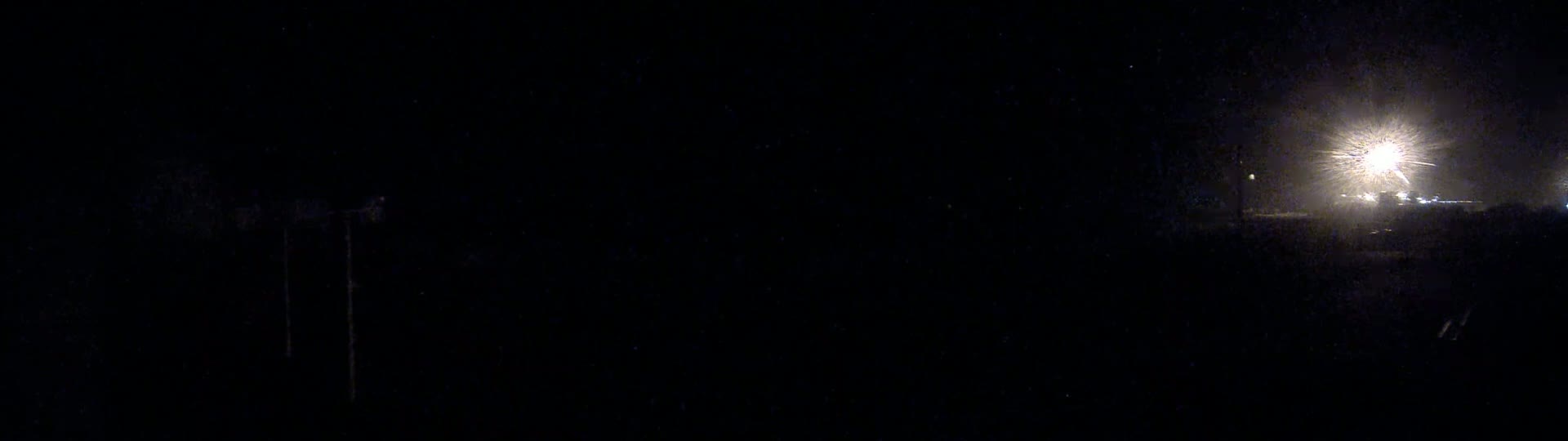 A nighttime, dark scene shows a bright light source in the distance, with a mostly dark foreground containing indistinct shapes.