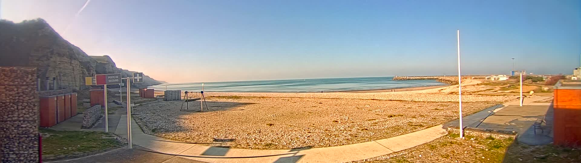 A sandy beach with a calm sea under a clear, bright sky stretches out before a rocky cliff, with a paved walkway and small buildings nearby.