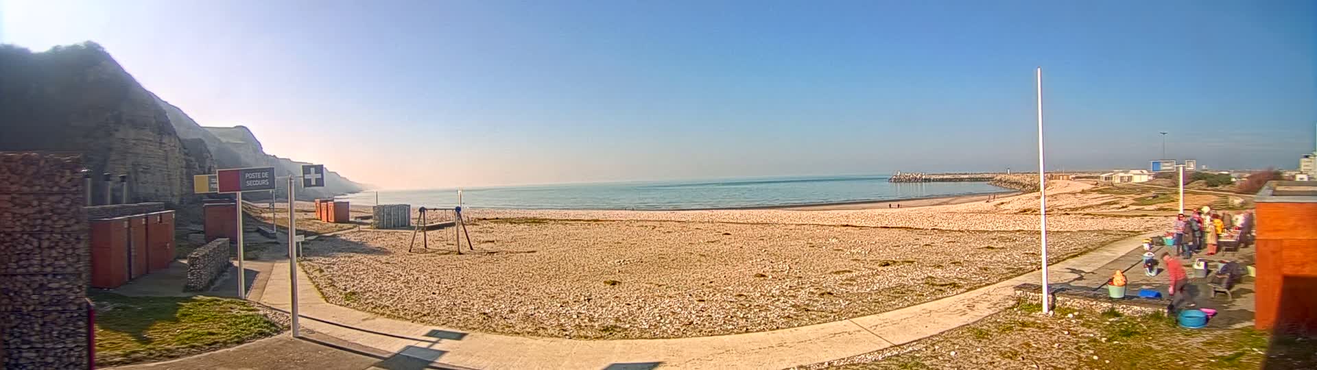 A sandy beach stretches out to a calm sea under a clear blue sky, backed by a rocky cliff face and a small group of people gathered near some buildings.