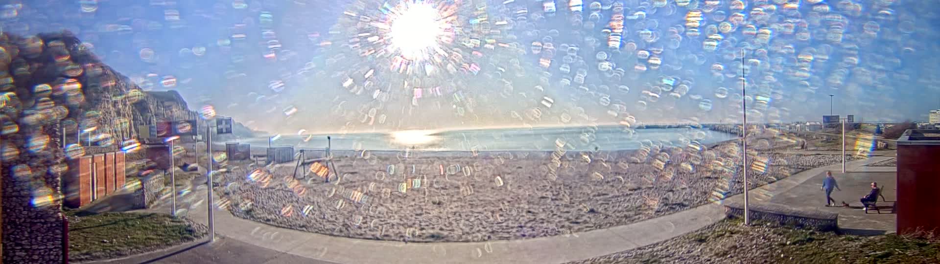 A beach scene on a sunny day is viewed through a distorted lens, showing a sandy beach, calm ocean water, a distant cliff, and people near a walkway.