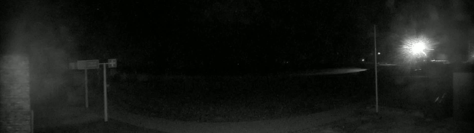 A nighttime, low-light image shows a dark, empty road with two tall posts and signs, and a bright light source in the distance.