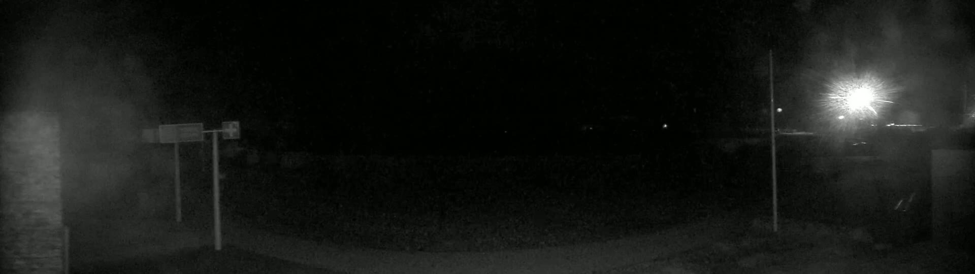 A nighttime, dimly lit outdoor scene shows two tall poles with rectangular signs, a bright light in the distance, and dark ground.