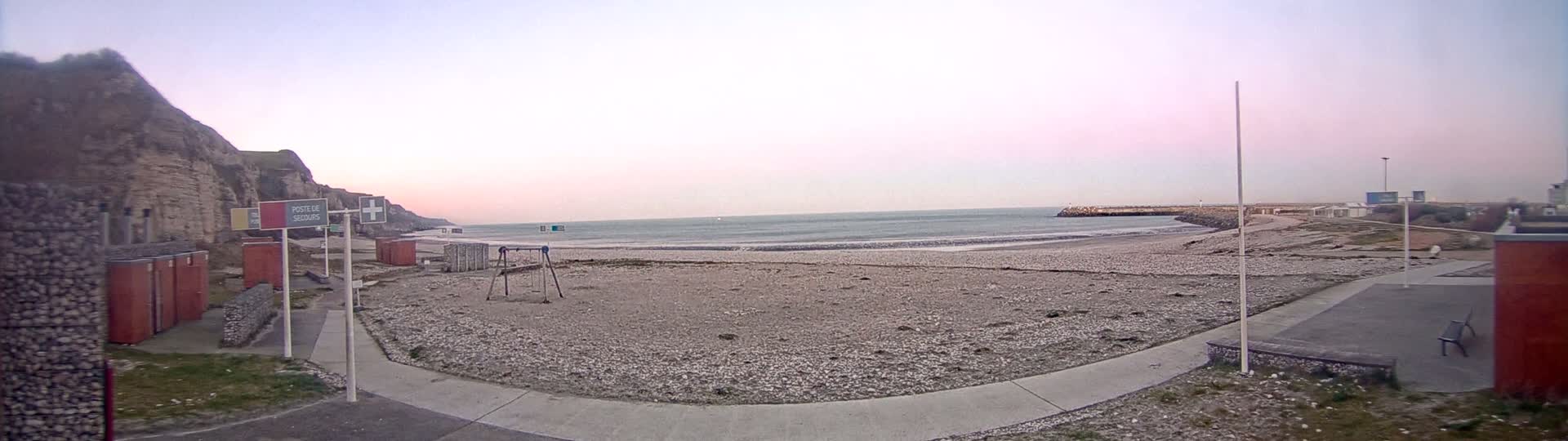 A pebble beach with a calm sea under a pale pink sky sits below a cliff face, with a small playground and changing rooms nearby.