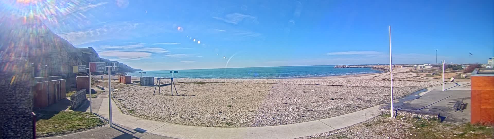 A mostly sunny day at a pebble beach with calm ocean waves, a stone cliff, and some beachside structures including a playground.