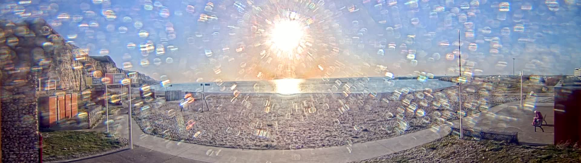 A beach scene with a bright sun shining over calm water, seen through a filter that creates a blurry, iridescent effect, with a rocky cliff in the background.