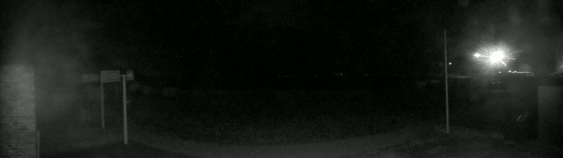 A dimly lit, nighttime outdoor scene shows a dark, empty area with two tall posts and a bright light source in the distance.