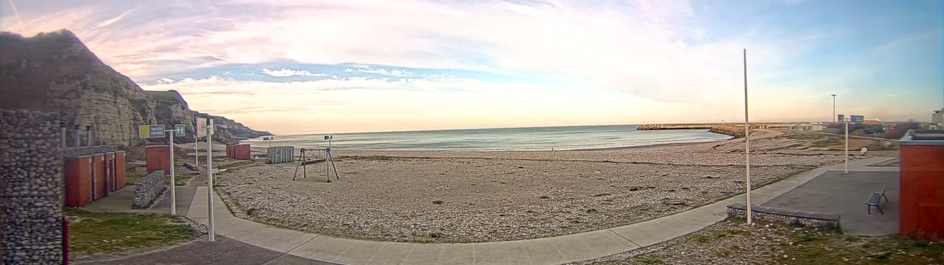 A mostly empty pebble beach is seen at sunset under a partly cloudy sky, with a rocky cliff to the left and a breakwater to the right.