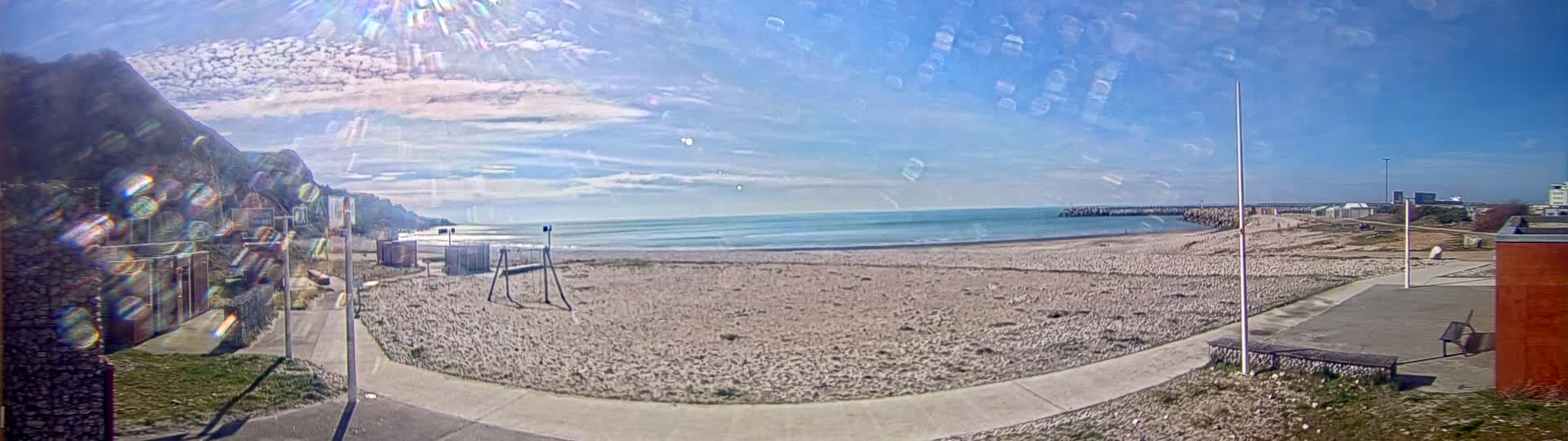 A mostly sunny day at a beach with a sandy shore, calm ocean, and a cliff in the background.