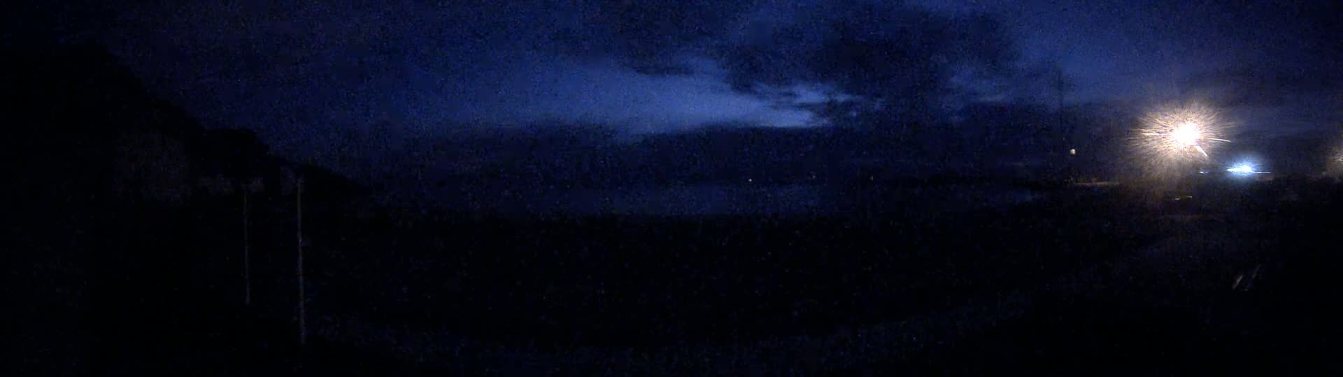 A dark, nighttime scene shows a cloudy sky illuminated by bright lights in the distance.