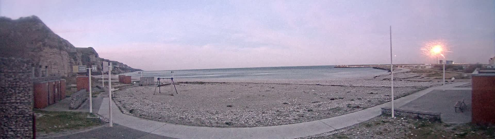 A pebble beach with a playground and a stone cliff in the background under a dusky sky with a firework display in the distance.