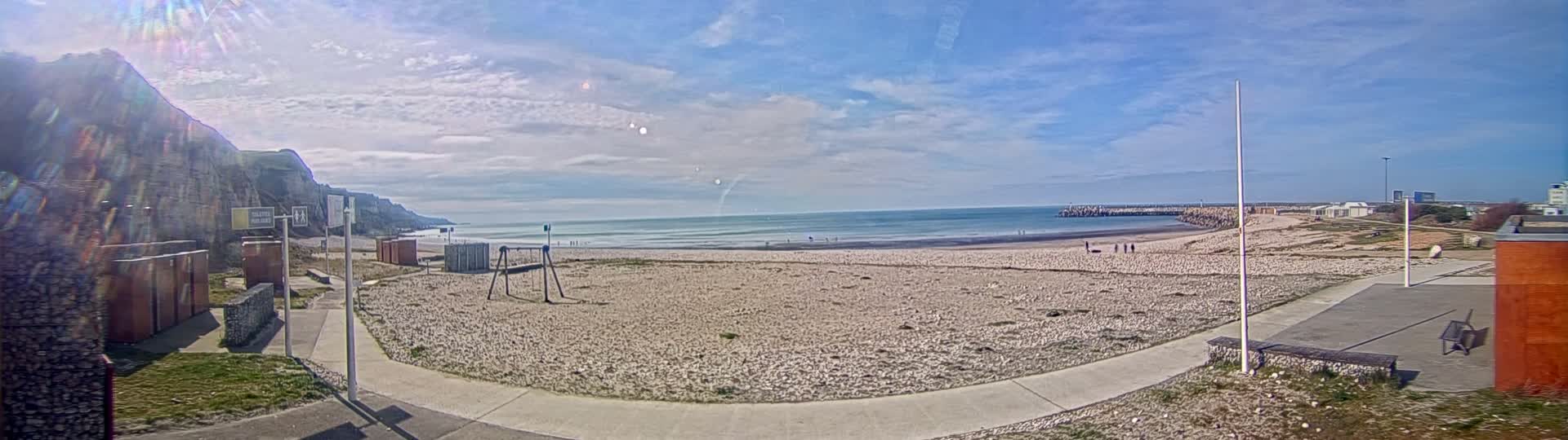 A mostly sunny day at a sandy beach, with a rocky cliff in the background and a few people scattered on the shore.