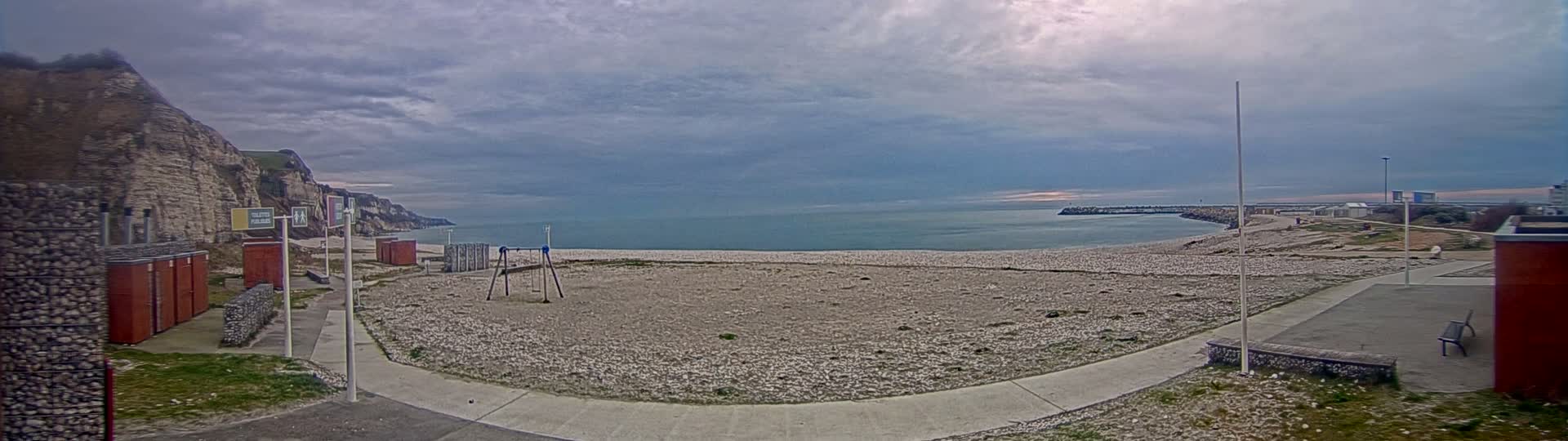 A mostly cloudy day reveals a pebble beach, a small playground, and a seaside walkway next to tall white cliffs, with a calm sea visible in the background.