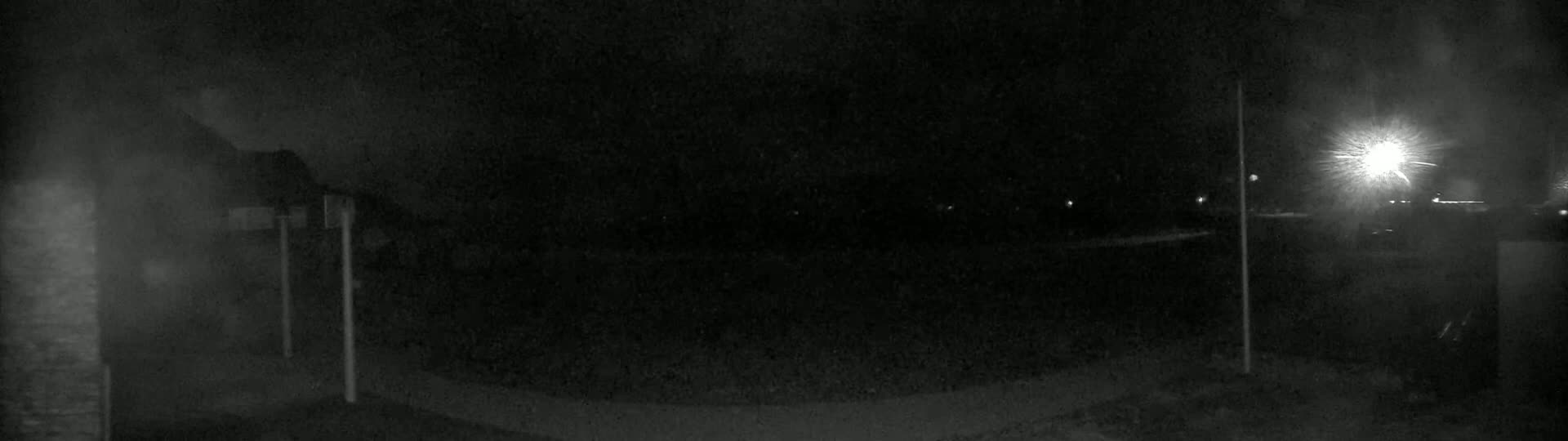 A nighttime, dimly lit outdoor scene shows a dark expanse of land with a few distant lights and a bright light source in the upper right corner, possibly a streetlight.