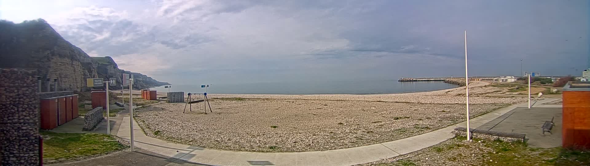 A mostly sunny day shows a pebble beach with a children's playground, restrooms, and a low sea wall, all situated next to a steep, rocky cliff overlooking a calm sea.