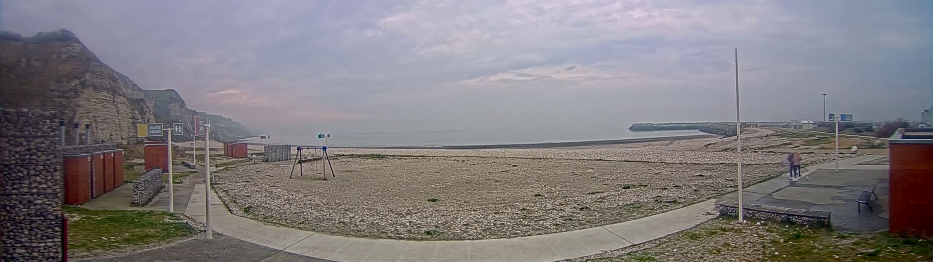 A mostly cloudy day reveals a pebble beach with a small playground,  adjacent to a rocky cliff and a calm sea.