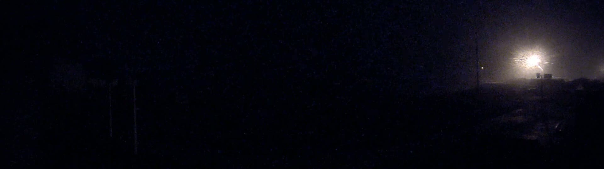 A bright light shines through the heavy nighttime rain, illuminating a dark, indistinct landscape.