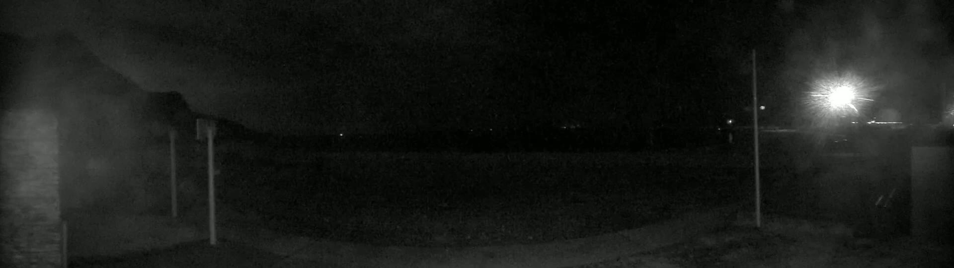 A nighttime, low-light view shows a dark, empty area with a bright light source in the distance and two tall, thin poles in the foreground.