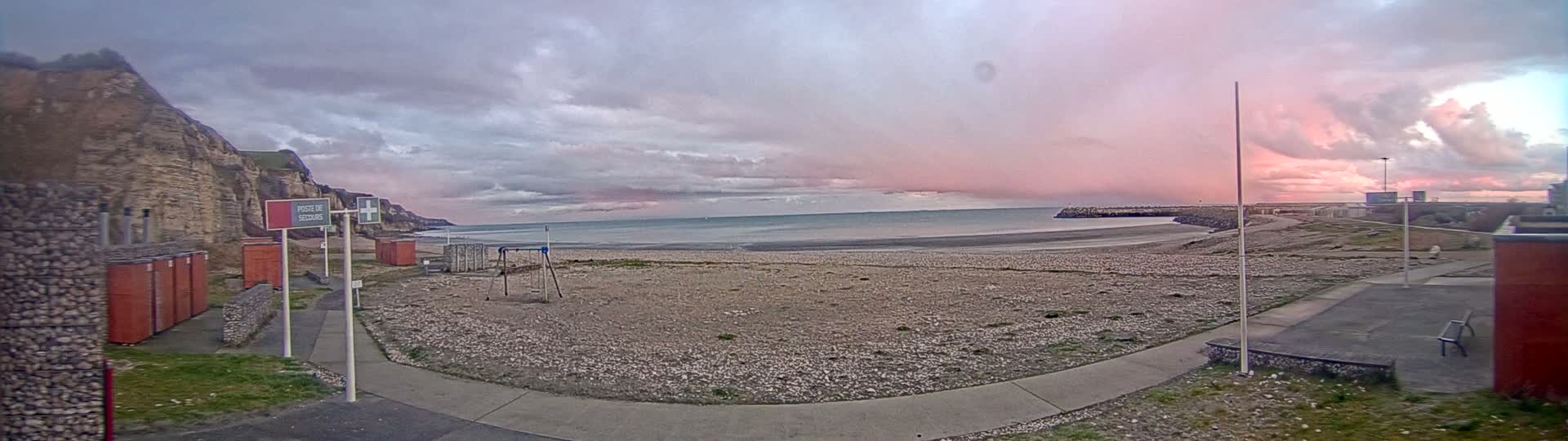 A pebble beach sits beneath a pink and grey cloudy sky, with a rocky cliff face to the left and a breakwater to the right.